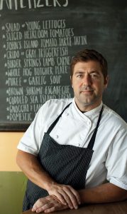 Chef Mike Lata posing in front of a chalkboard menu.