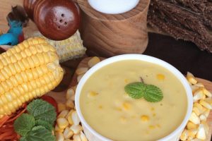 A bowl of shrimp and corn bisque surrounded by corn and other vegetables.