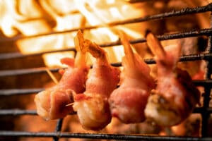 A row of wild-caught American shrimp wrapped in bacon being cooked on a fiery grill.