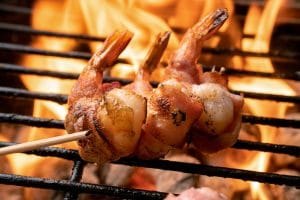 A row of wild-caught American shrimp wrapped in bacon being cooked on a fiery grill.