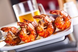 A plate of grilled shrimp on a skewer with a glass of beer.